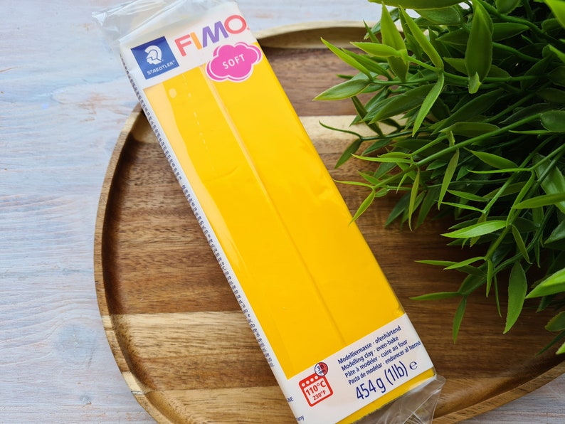 Puede incluir: Un paquete de arcilla de modelado amarilla FIMO SOFT, que pesa 454 gramos, se muestra en una bandeja de madera. El paquete tiene los logotipos STAEDTLER y FIMO SOFT, con texto que indica que es arcilla para hornear. La temperatura se muestra en grados Celsius.