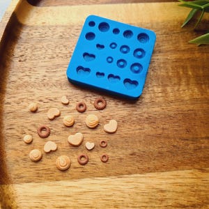 Op de afbeelding: Een blauwe siliconen mal met verschillende vormen om kleine lekkernijen te maken, zoals harten en cirkels. De mal ligt op een houten oppervlak met verschillende kleine, bruine, hartvormige en ronde lekkernijen.