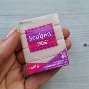 May include: A package of Sculpey Premo polymer clay, 2 oz (57 g), in a light beige color. The package has a purple and pink label with the brand name and product information. The clay is oven-bake and is held in a hand.