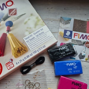 May include: A FIMO Leather Effect Tassel Kit with four 25 gram blocks of polymer clay in black, blue, pink, and a small package of black string. The kit includes instructions for making tassels and comes with four silver jump rings. The box is labelled "Tassel Kit - Quasten Set - Kit Tassels - Set Borlas" and includes the text "4 x 25 g (0.88 oz) 100 g (3.5 oz)".