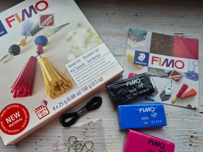 May include: A FIMO Leather Effect Tassel Kit with four 25 gram blocks of polymer clay in black, blue, pink, and a small package of black string. The kit includes instructions for making tassels and comes with four silver jump rings. The box is labelled "Tassel Kit - Quasten Set - Kit Tassels - Set Borlas" and includes the text "4 x 25 g (0.88 oz) 100 g (3.5 oz)".