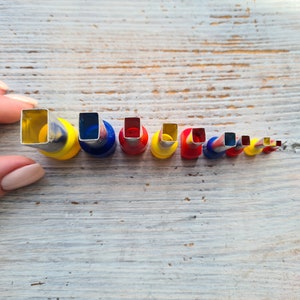 May include: A collection of square-shaped metal craft punches in various sizes, held by a hand. The punches have colorful bases of yellow, blue, and red. The sizes decrease from left to right.