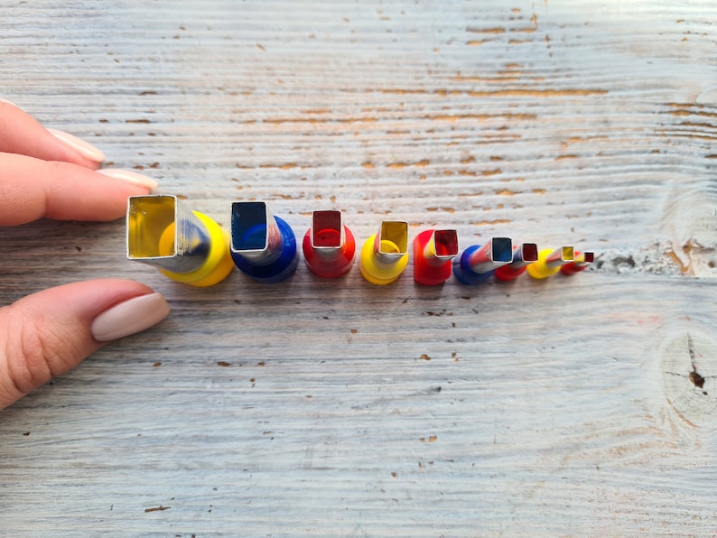 May include: A collection of square-shaped metal craft punches in various sizes, held by a hand. The punches have colorful bases of yellow, blue, and red. The sizes decrease from left to right.