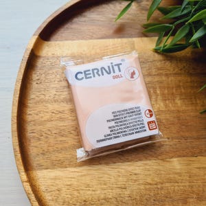 May include: A package of Cernit Doll polymer clay in a skin-tone color. The package has text in multiple languages, including "Skin Effect Polymer Clay." The clay is displayed on a wooden tray with green foliage.
