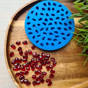 Könnte beinhalten: Eine blaue Silikonform mit mehreren kleinen, unregelmäßigen Formen, die an Edelsteine erinnern. Um die Form herum sind zahlreiche kleine, rote, durchscheinende, edelsteinartige Objekte verstreut.