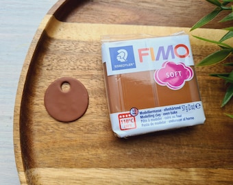 May include: A package of FIMO Soft modeling clay in brown. The package has the text "FIMO Soft" and "Modelliermasse ofenhärtend 57g (2 oz) e" on it. The package is sitting on a wooden surface with a brown clay circle.