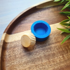 May include: A blue silicone cupcake mold with a fluted edge sits on a wooden surface next to a small, round, wooden cupcake liner.