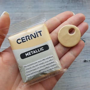 Könnte beinhalten: Eine Packung goldfarbener, metallischer Polymer-Knete mit dem Markennamen "CERNIT" auf der Vorderseite. Die Packung enthält den Text "Metallic" und Anweisungen in mehreren Sprachen.