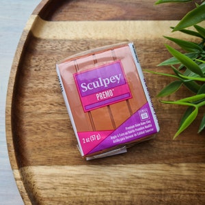 Könnte beinhalten: Eine 57 Gramm Packung Sculpey Premo Backofen-Modelliermasse. Die Verpackung ist rosa und lila mit dem Sculpey-Logo und den Worten "Premium Backofen-Modelliermasse".