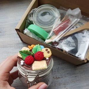 Puede incluir: Un tarro de vidrio con tapa lleno de crema de chocolate cubierta con caramelos de colores, galletas y fresas. El tarro está sobre una superficie de madera.