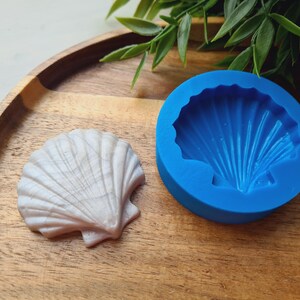 Könnte beinhalten: Eine leuchtend blaue, kreisförmige Silikonform mit einer vertieften Muschelform mit strahlenförmigen Rillen, platziert neben einem fertigen, perlweiß schimmernden Muschelabguss, der dieselbe detaillierte, fächerartige Rippenstruktur aufweist.