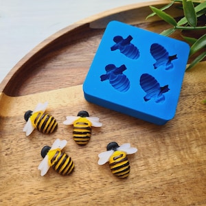 May include: A wooden tray holds three small bee figurines with black and yellow striped bodies and white wings. A blue silicone mold with four bee-shaped cavities sits nearby. Green leaves are in the background.