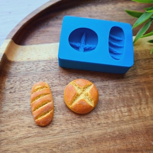 May include: A blue silicone mold with two cavities, one round and one oblong, next to two miniature bread replicas. One is a round roll with a cross design, and the other is an oblong loaf. The items are on a wooden tray.