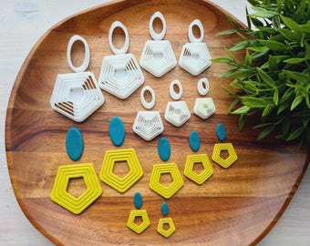 May include: A collection of handmade polymer clay earrings in various sizes and shapes, including white geometric designs, teal ovals, and yellow pentagons. The earrings are arranged on a wooden tray with a green plant in the background.
