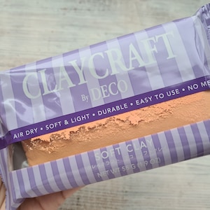 Può includere: Un pacchetto di argilla ad asciugatura all'aria etichettato "Claycraft by Deco". La confezione è viola e bianca con strisce e il testo "Air Dry, Soft & Light, Durable, Easy to Use, No Mess". L'argilla è di colore arancio chiaro e la confezione dice "Soft Clay" e "Net Wt 55 g (1.9 oz)".