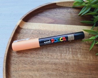 May include: A peach-colored Uni Posca paint marker with black lettering and a black cap. The marker is on a wooden surface.