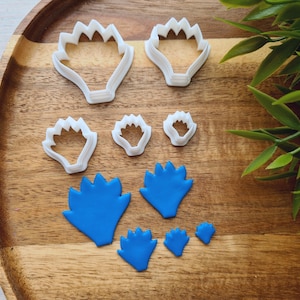 Puede incluir: Un conjunto de ocho cortadores de galletas de plástico blanco en varias formas de flores sobre una superficie de madera. Los cortadores están dispuestos en un patrón con el cortador más grande en el centro. También hay cinco formas de flores de fondant azul en la superficie.