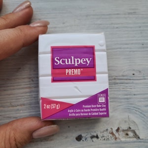 May include: A white pack of Sculpey Premo polymer clay, 57 g. The package has a pink and purple label with the brand name and product information. The clay is premium oven-bake clay.
