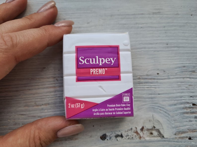 May include: A white pack of Sculpey Premo polymer clay, 57 g. The package has a pink and purple label with the brand name and product information. The clay is premium oven-bake clay.