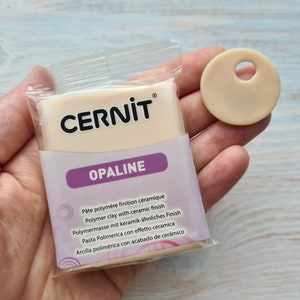 Może przedstawiać: Opakowanie białej gliny polimerowej z ceramicznym wykończeniem. Opakowanie jest oznaczone "CERNIT OPALINE" i zawiera wielojęzyczne opisy produktu. Pokazano również mały, okrągły, biały krążek gliny.