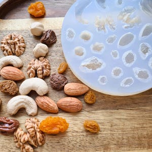 May include: A silicone mold with multiple cavities shaped like nuts and dried fruit. The mold is on a wooden surface with a variety of nuts and dried fruit scattered around it.