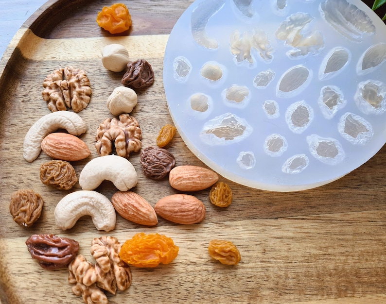 May include: A silicone mold with multiple cavities shaped like nuts and dried fruit. The mold is on a wooden surface with a variety of nuts and dried fruit scattered around it.