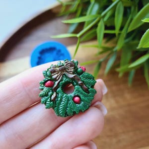 Puede incluir: Una corona de Navidad verde con un lazo marr&oacute;n y bayas rojas. La corona est&aacute; hecha de pl&aacute;stico y tiene un di&aacute;metro de unos 5 cm.