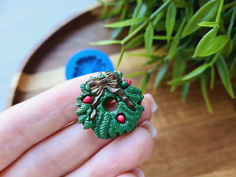 Puede incluir: Una corona de Navidad verde con un lazo marr&oacute;n y bayas rojas. La corona est&aacute; hecha de pl&aacute;stico y tiene un di&aacute;metro de unos 5 cm.