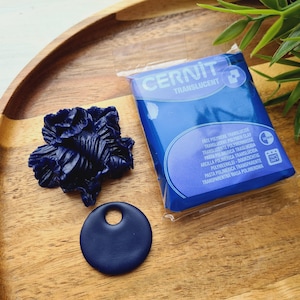 May include: A wooden tray holds a dark blue polymer clay flower, a round pendant with a hole, and a package of Cernit Translucent polymer clay. The package is blue with white text.