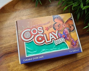 Può includere: Una scatola rettangolare di Cosclay Deco, un materiale modellabile in plastica flessibile. La confezione presenta illustrazioni di farfalle arancioni e viola, il testo 'Cosclay Deco', 'The Fantastic Flexible Plastic' e 'Curable Oven Safe'. Pesa 453g.