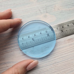 May include: A clear, round, glass disc measuring 2 inches in diameter. The disc is being measured with a ruler.