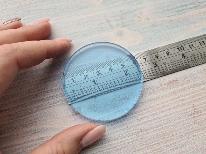 May include: A clear, round, glass disc measuring 2 inches in diameter. The disc is being measured with a ruler.