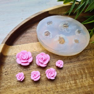 May include: A collection of pink rose-shaped embellishments and a silicone mold. The roses vary in size and are arranged on a wooden tray. The mold is oval-shaped with multiple flower impressions.