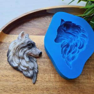 Pode incluir: Uma escultura detalhada da cabeça de um lobo em tons de cinza, preto e branco, ao lado de um molde de silicone azul do mesmo desenho. A escultura está em uma bandeja de madeira, com folhagem verde ao fundo. A escultura do lobo tem cerca de 10 cm de comprimento.