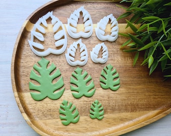 May include: A set of white plastic cookie cutters shaped like monstera leaves, and a set of green fondant monstera leaves on a wooden tray.