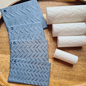 May include: A selection of textured, grey-blue square tiles and white cylindrical objects, all featuring a diamond plate pattern. The items are displayed on a wooden tray, offering a contrast in colour and texture.