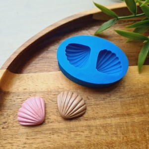 May include: A bright blue oval silicone mold featuring two recessed seashell shapes, accompanied by one small pink seashell-shaped piece and one small tan seashell-shaped piece, each displaying detailed radial ridges on their surfaces.