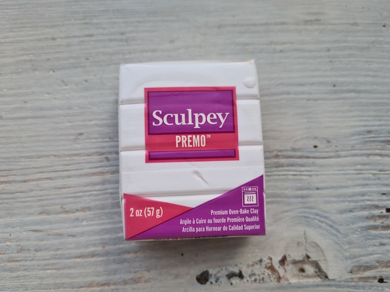 May include: A package of white Sculpey Premo oven-bake clay, 57 g. The packaging features a purple and pink label with the brand name and product details. The package also includes text in French and Spanish.