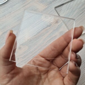 May include: A clear glass square, held in a hand, with a smooth surface.