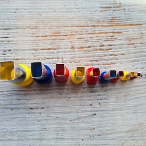 May include: A set of colorful metal craft punches with square openings, arranged in a row from largest to smallest. The punches have yellow, red, and blue accents and are set against a weathered, light-colored wooden surface.