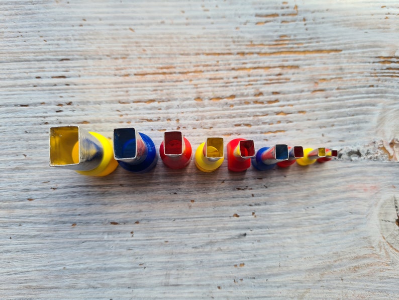 May include: A set of colorful metal craft punches with square openings, arranged in a row from largest to smallest. The punches have yellow, red, and blue accents and are set against a weathered, light-colored wooden surface.