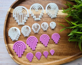 May include: A set of 10 white and pink plastic cookie cutters in the shape of a stylized rainbow arch. The cutters are arranged on a wooden surface.