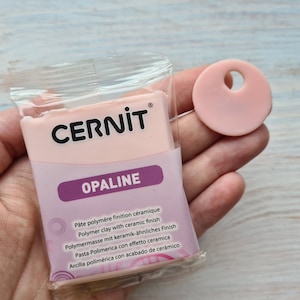 Może przedstawiać: Opakowanie gliny polimerowej Cernit Opaline i mała, okrągła, różowa zawieszka z otworem. Opakowanie gliny jest różowe z czarnym napisem "Cernit" i białym słowem "OPALINE".