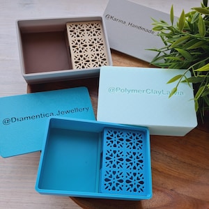 May include: A selection of jewellery boxes and display cards in various colours. The boxes are rectangular with a decorative insert. The display cards have the text "@Dianmentica.Jewellery" and "@PolymerClayLavia". The boxes are in shades of blue, grey, and brown.