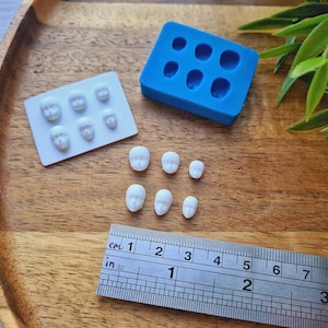May include: A wooden tray holds a blue silicone mould, a white mould with face shapes, and several small, white face-shaped figurines. A ruler shows measurements in inches and centimetres. The figurines vary in size, with the largest being about 2 cm.