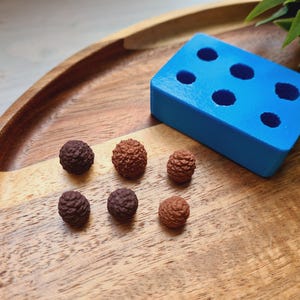 May include: A blue silicone mould with six round cavities, sitting on a wooden tray. There are six small brown and tan pinecone-shaped objects on the tray.