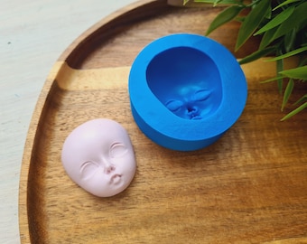 Könnte beinhalten: Eine blaue Silikonform zum Herstellen eines Baby-Puppenkopfes. Die Form befindet sich auf einer Holzoberfläche neben einem rosafarbenen Puppenkopf.