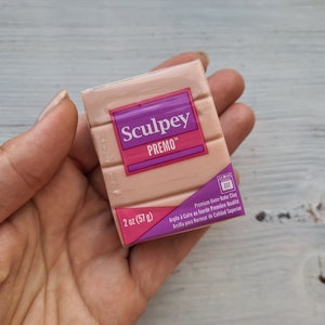 Puede incluir: Un bloque de arcilla para hornear Sculpey Premo de 2 onzas (57 gramos). La arcilla es de color marrón claro y está envasada en una caja de color morado y blanco.