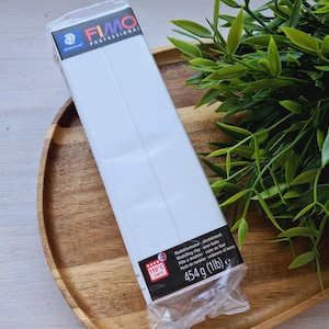 Pode incluir: Uma embalagem branca de argila para modelar FIMO Professional. A embalagem está sobre uma superfície de madeira e tem um rótulo vermelho e branco com o texto "110°C 230°F" e "454 g (1 lb)".