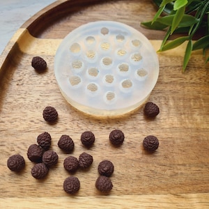 Pode incluir: Um molde de silicone transparente com 24 cavidades redondas, perfeito para fazer pequenas artesanato de resina. O molde est&aacute; sobre uma superf&iacute;cie de madeira com v&aacute;rias pequenas pinhas de resina marrom espalhadas ao redor.